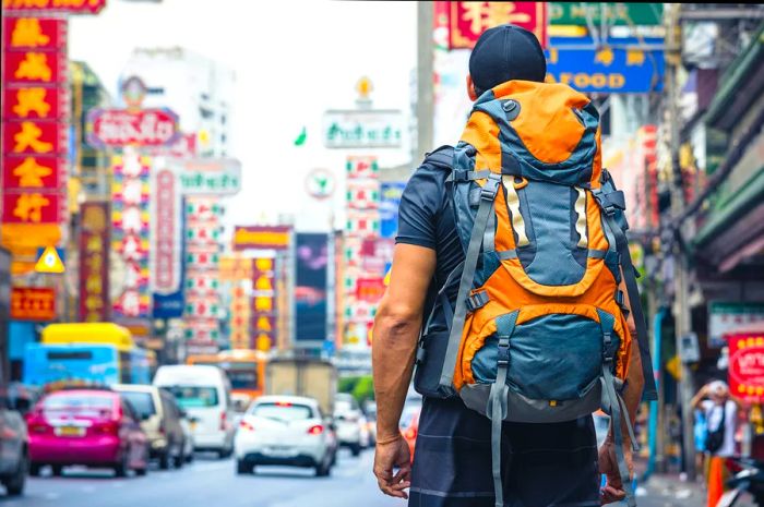 A traveler with a bright yellow backpack stands on a bustling street in Bangkok, gazing at the traffic and vibrant neon signs.