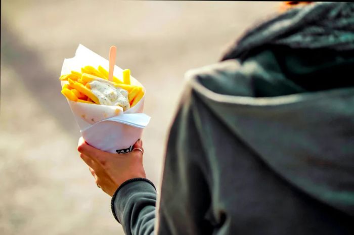 A person strolls down a street enjoying fries from a paper cone