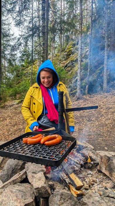 A woman relaxes by a campfire in the woods, grilling sausages.