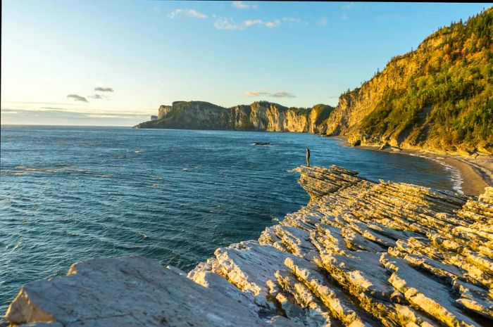 A person stands on the rocky shoreline backed by cliffs