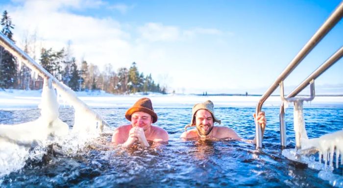 Two men wearing hats smile broadly as they immerse themselves in a frozen pool after warming up in a sauna