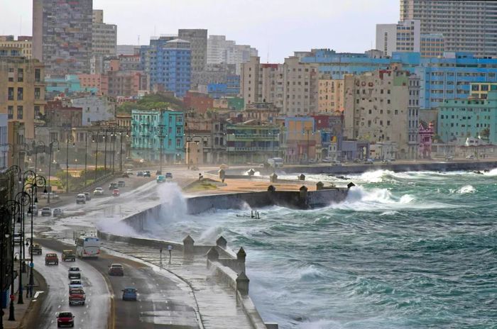 Powerful waves crash against the shore, sweeping over the road as cars pass by.