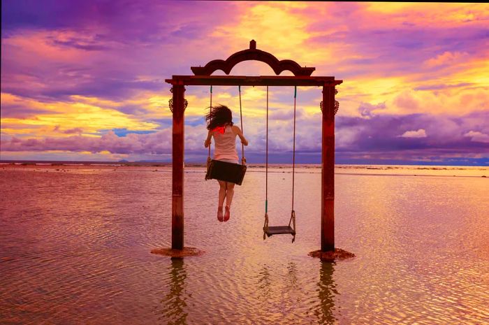 A woman joyfully swings high on a swing set in the ocean's shallows as the sun sets, painting the sky in shades of pink.