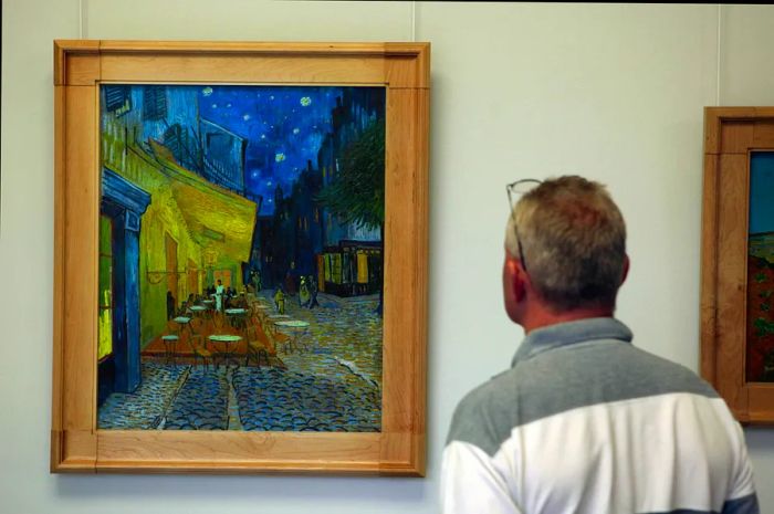 A man stands admiring a framed painting depicting people outside a cafe at night.