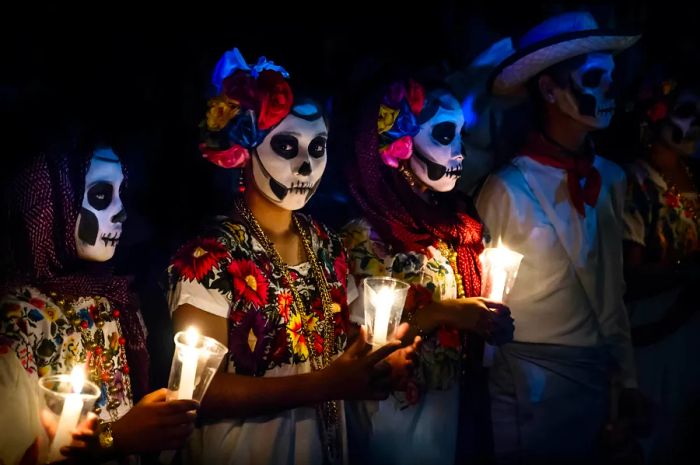 Three women in Catrina costumes and a man dressed in white cowboy attire, adorned with skull makeup and holding candles at a Día de Muertos parade