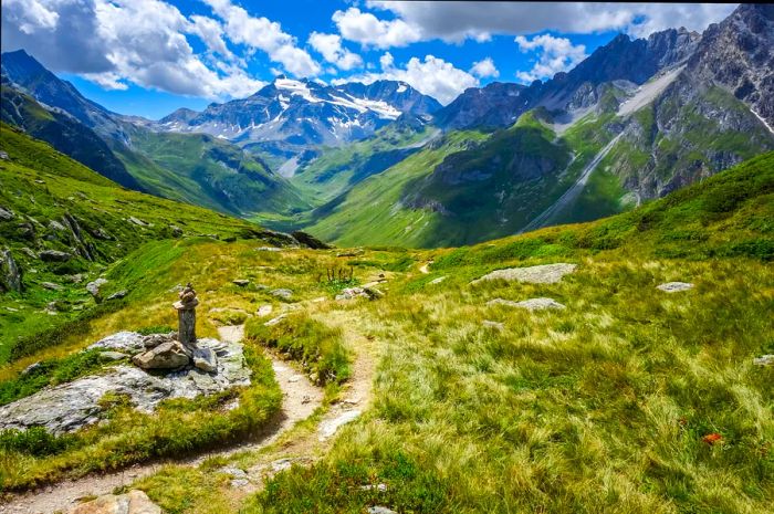 A rugged path meanders through a rocky, grassy alpine terrain, with snow-capped peaks visible in the distance.