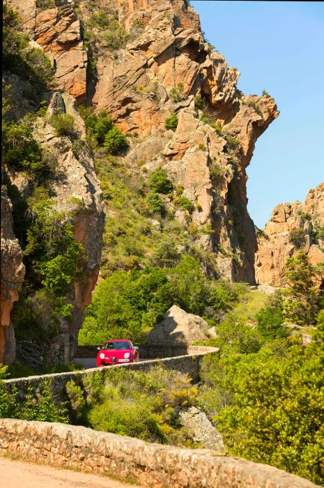 A red car navigates a bend on a narrow road hugging a towering cliff face.