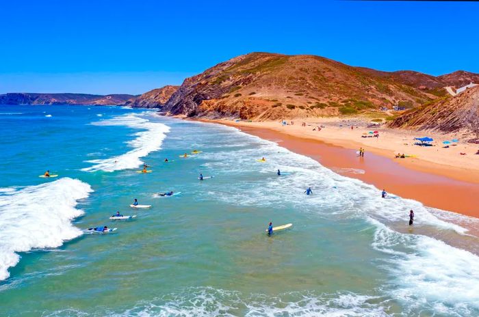 An aerial view captures surfers riding the waves along a rugged red coastline in Portugal.