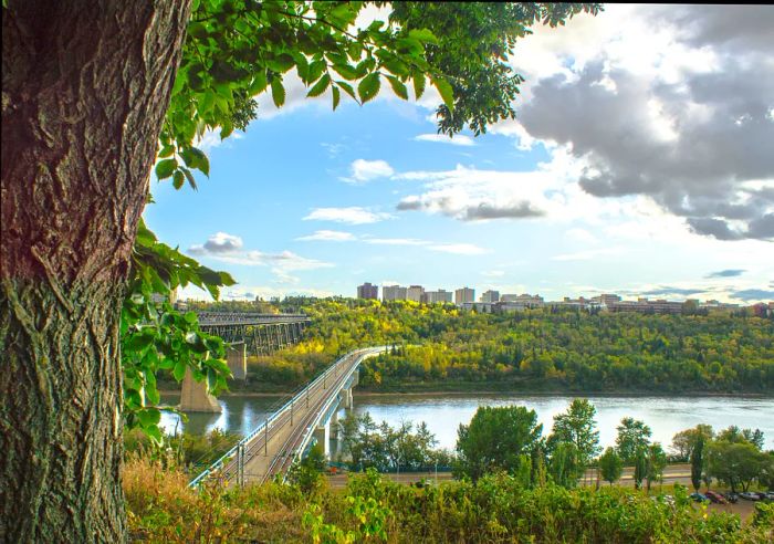 A scenic afternoon view of the North Saskatchewan River Valley alongside downtown Edmonton, the capital of Alberta. The train bridge linking the city's east and west sides is also visible. It’s the start of autumn, with leaves beginning to change to yellow, orange, and red hues.