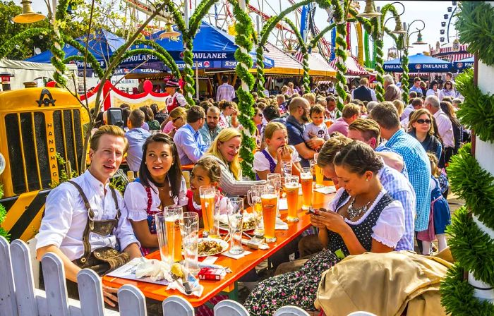 People in traditional attire gather around long wooden tables in a bustling beer garden.