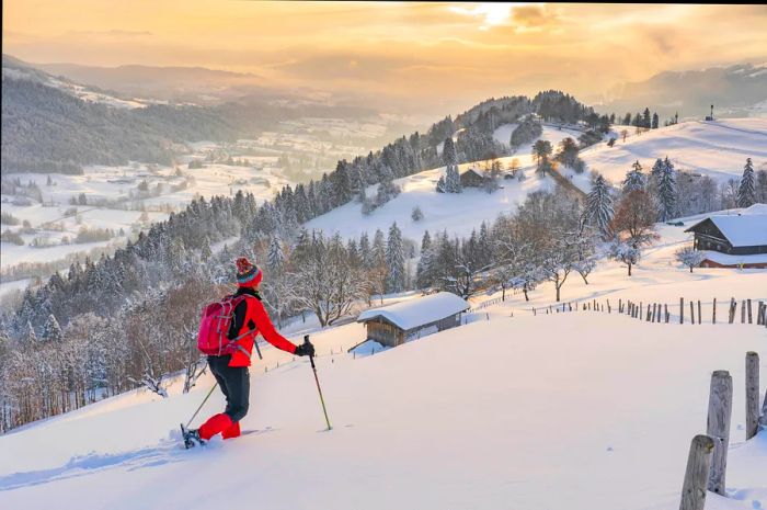 A person fully equipped for skiing snowshoes through deep snow in a mountainous area as the sun sets.