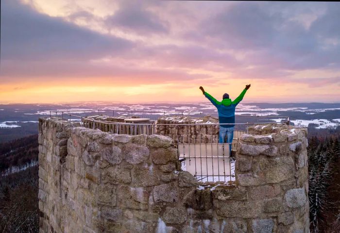 A man clad in winter attire stands atop a crumbling castle tower, gazing out over the snow-covered landscape.