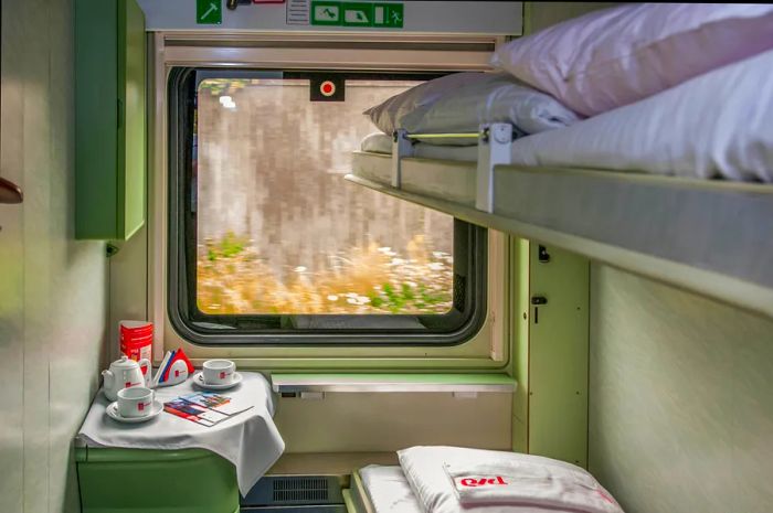 Inside a sleeper train cabin, featuring two compact bunks on the right wall and a window showcasing the scenic views outside