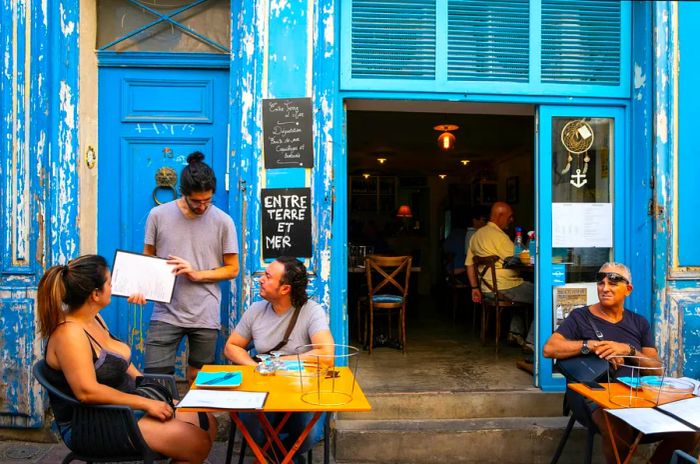 A waiter serves a tourist couple on the terrace of a bistro in the vibrant Le Panier neighborhood, one of Marseille’s most lively and tourist-friendly areas.