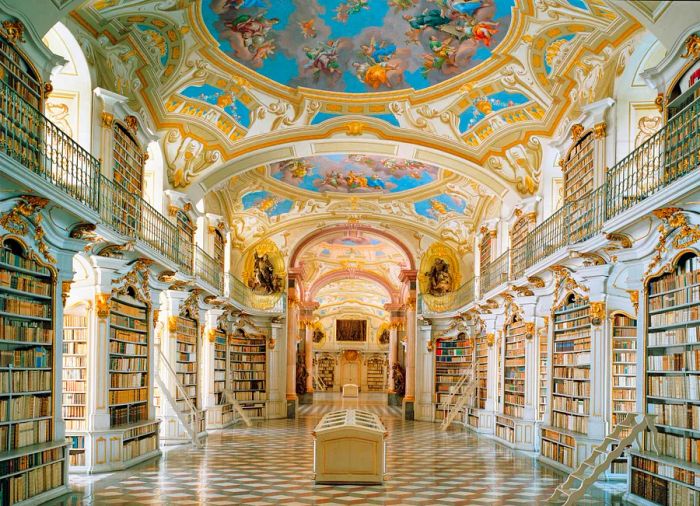 A grand hall supported by rectangular pillars that also function as bookcases, each adorned with white and gilded baroque details. A balcony encircles the upper level, while the ceiling above features a striking combination of white, blue, and gold vaults, enhancing the architectural elegance.