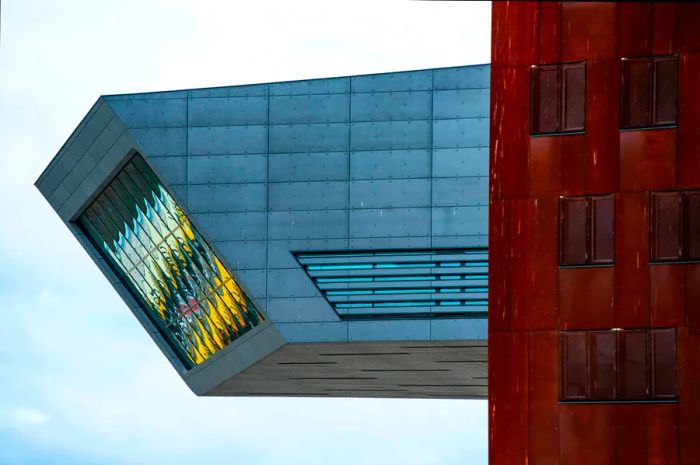 A sleek modernist cantilevered wing of the library at the Vienna University of Economics extends from a brown structure, culminating in a rectangular glass wall that tilts downward, reflecting a spectrum of colors from light blue to yellow.