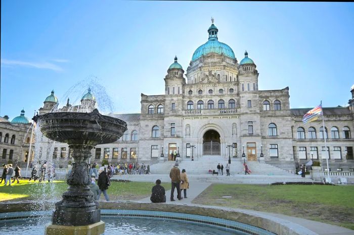 A crowd gathers around a fountain outside the Legislative Assembly Parliament Building of British Columbia in Victoria on a sunny day.