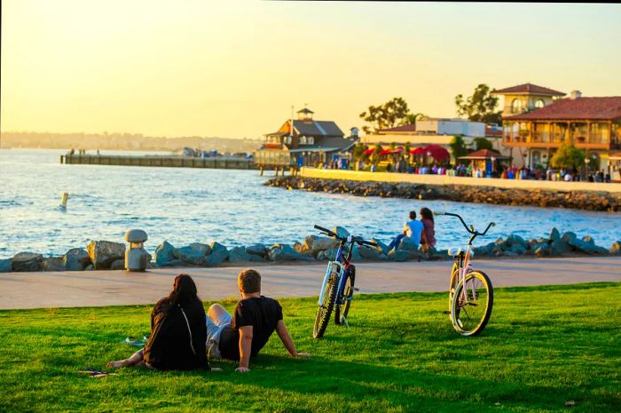 People relax on the grass by the waterfront as the sun sets