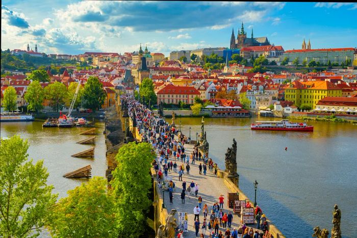 Summer visitors fill a bridge over a river leading to a castle atop a hill on a sunny day