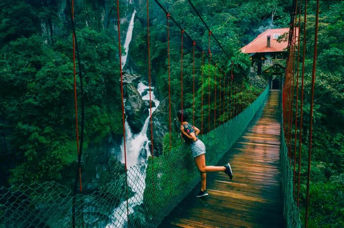 A woman in shorts stands on a suspension bridge, gazing at a waterfall in Ecuador.