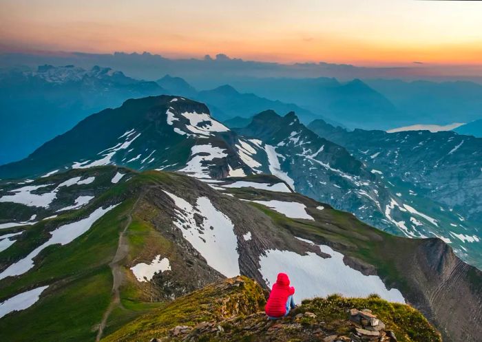 A solitary hiker gazes at the sunset over a tranquil lake nestled in the mountains