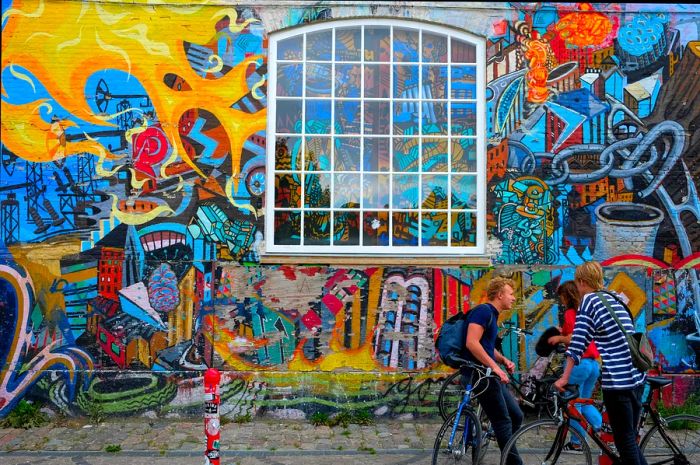 Groups of young people with bikes gather in front of a vibrant graffiti wall in Christiania, Copenhagen