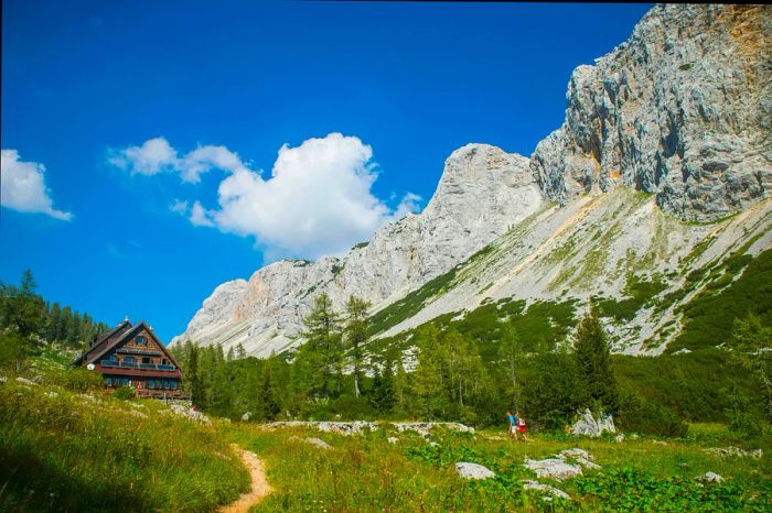 Two hikers traverse a trail leading to a rustic cabin amidst a rugged landscape