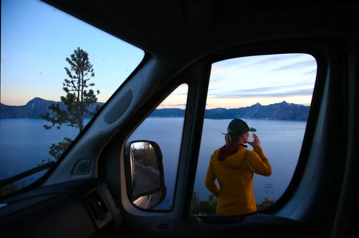 A young woman savors a drink beside her car on the shore of Crater Lake, Crater Lake National Park, Oregon, USA