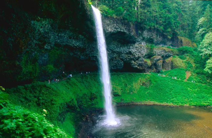 A waterfall cascades dramatically into a pool, with hikers behind it appearing as tiny figures.