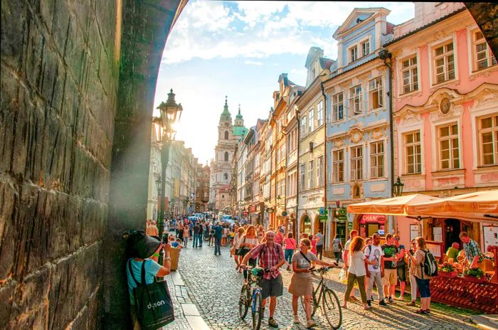 At sunset in summertime, people stroll and cycle along a bustling cobblestone street in Prague.