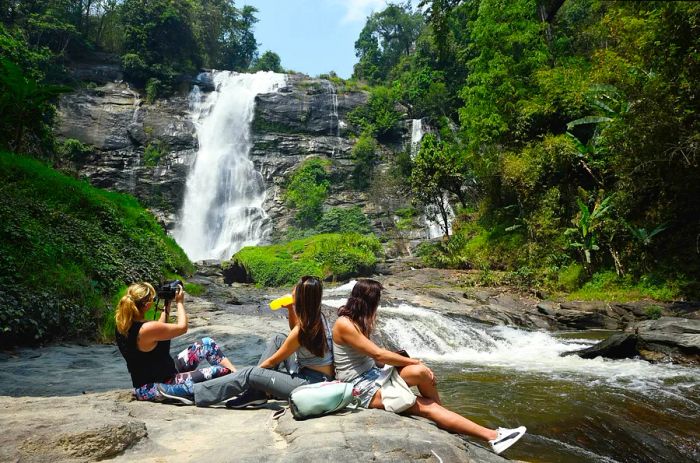 Three individuals relax on a rock beside a pool at a rushing waterfall