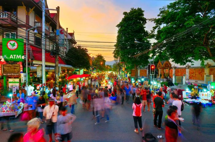 A bustling night market stretches along one of the main roads in Chiang Mai's old city, with a stream of people (blurred to indicate they are in motion) strolling alongside the stalls.