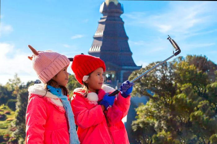 Two young children, bundled in coats and furry hats, pose for a selfie in Chiang Mai with a temple stupa in the background.