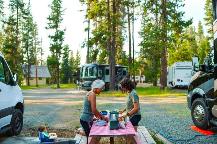 Two women enjoy cooking at a picnic table next to an RV parked in a picturesque, tree-lined campground