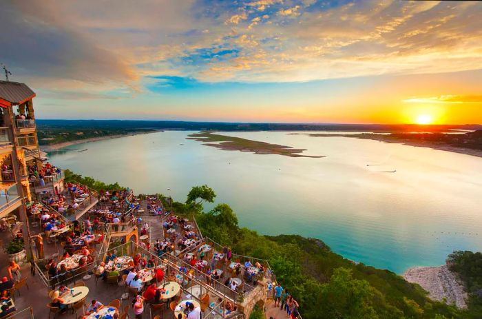 Diners enjoy a meal on a terrace by a lakeside during sunset.