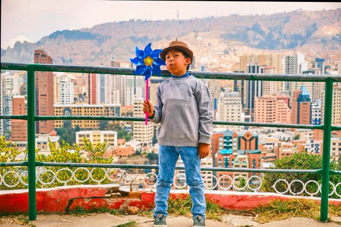 A boy in a hat joyfully spins a pinwheel in the city.