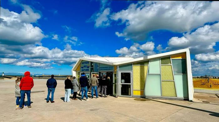 A queue of customers forms outside a seaside kiosk offering food.