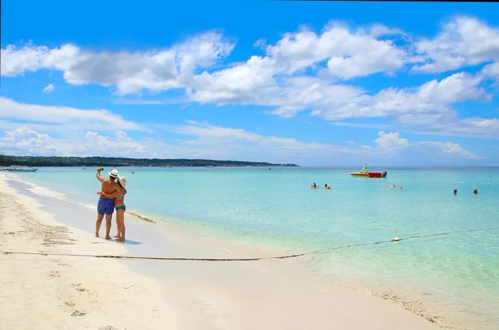 A distant view of two people taking a selfie on Negril Beach while others enjoy a swim