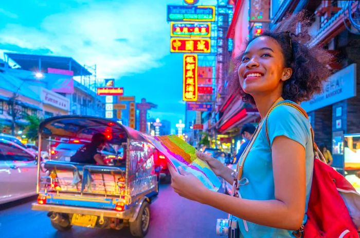 A cheerful young woman is seen holding a map on a bustling street in Bangkok, Thailand.