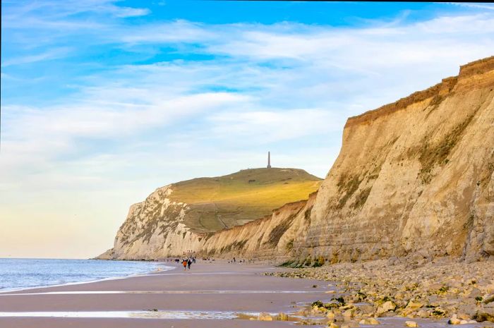 Visitors stroll along a beach next to a towering cliff crowned with an obelisk.