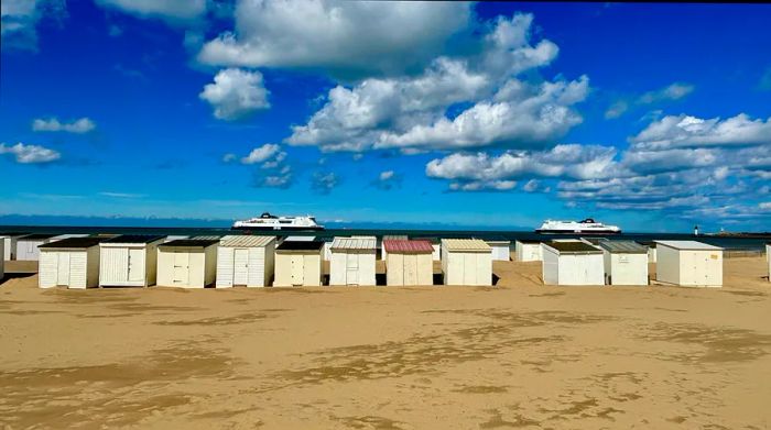 A pair of ferries navigating the sea, with a line of beach huts in the foreground