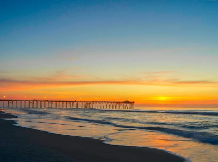 A wooden fishing pier extends into the ocean as the sun rises, casting an orange glow across the sky