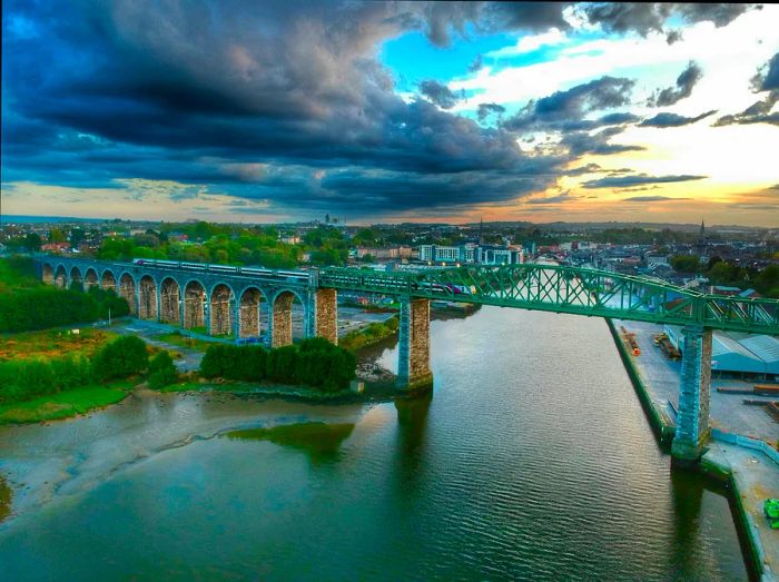 A train traverses a viaduct under a dramatic, cloud-filled sky