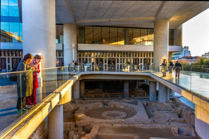 The entrance to the Acropolis Museum in Athens, featuring two women gazing at the excavated site from a bridge, Greece