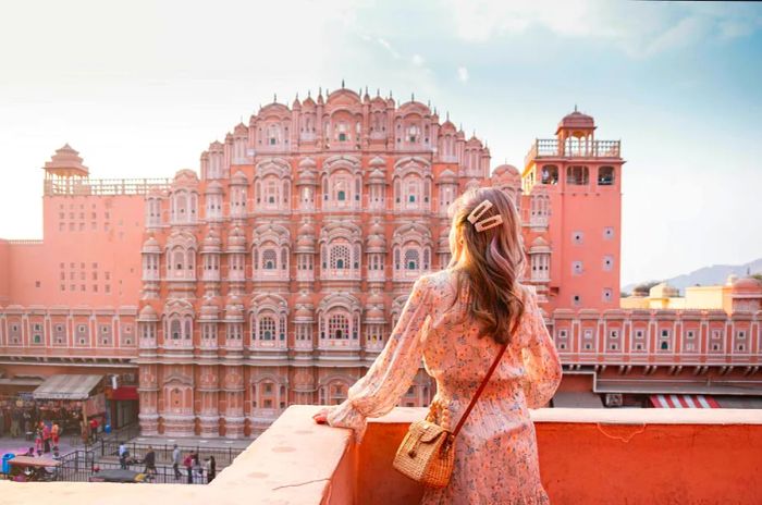 A woman stands on a rooftop gazing at a vast pink structure adorned with hundreds of windows.