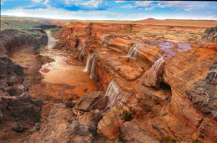 A series of small cascades pour into a river from a reddish-brown rocky cliff