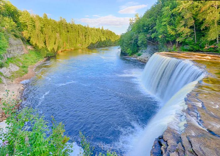 A sweeping waterfall cascades into a river flanked by lush woodlands