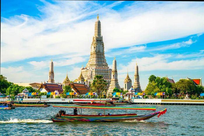 The Wat Arun temple at dawn with a boat gliding by on a sunny day in Bangkok, Thailand.