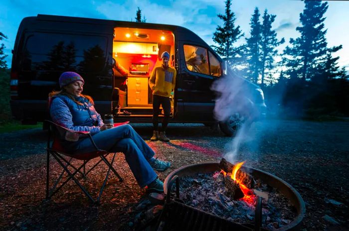 Two individuals enjoying a campfire outside their camper van at dusk in Denali National Park.