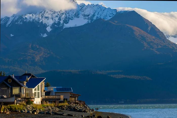 Two individuals stroll along the coastline, their shadows dwarfed by the towering mountains above them.